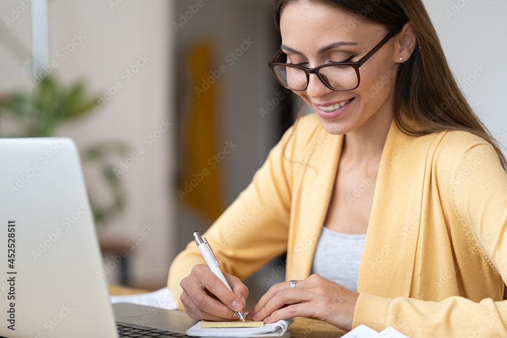 Young student woman studying from home use laptop computer watch educational video or e-learning courses making notes in notebook. Female entrepreneur passing certificate training learning online Young student woman studying from home use laptop computer watch educational video or e-learning courses making notes in notebook. Female entrepreneur passing certificate training learning online