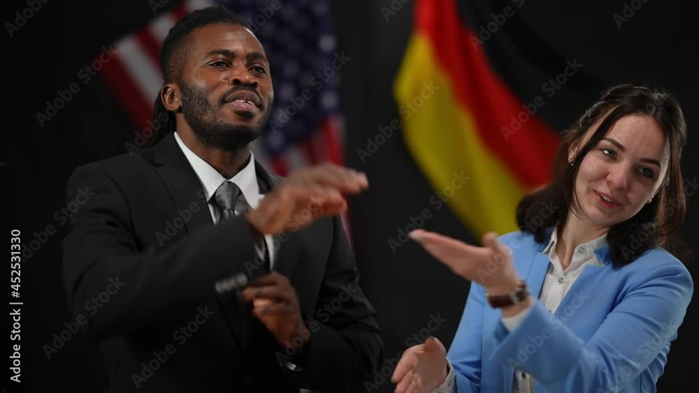 Clapping Caucasian woman inviting African American man to the stage in camera flashes at press conference. Positive female and male diplomats politicians greeting shaking hands smiling Clapping Caucasian woman inviting African American man to the stage in camera flashes at press conference. Positive female and male diplomats politicians greeting shaking hands smiling