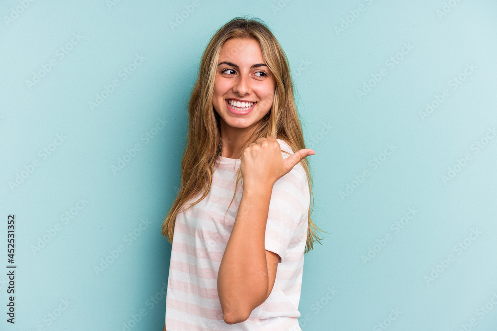 Young caucasian blonde woman isolated on blue background points with thumb finger away, laughing and carefree. Young caucasian blonde woman isolated on blue background points with thumb finger away, laughing and carefree.