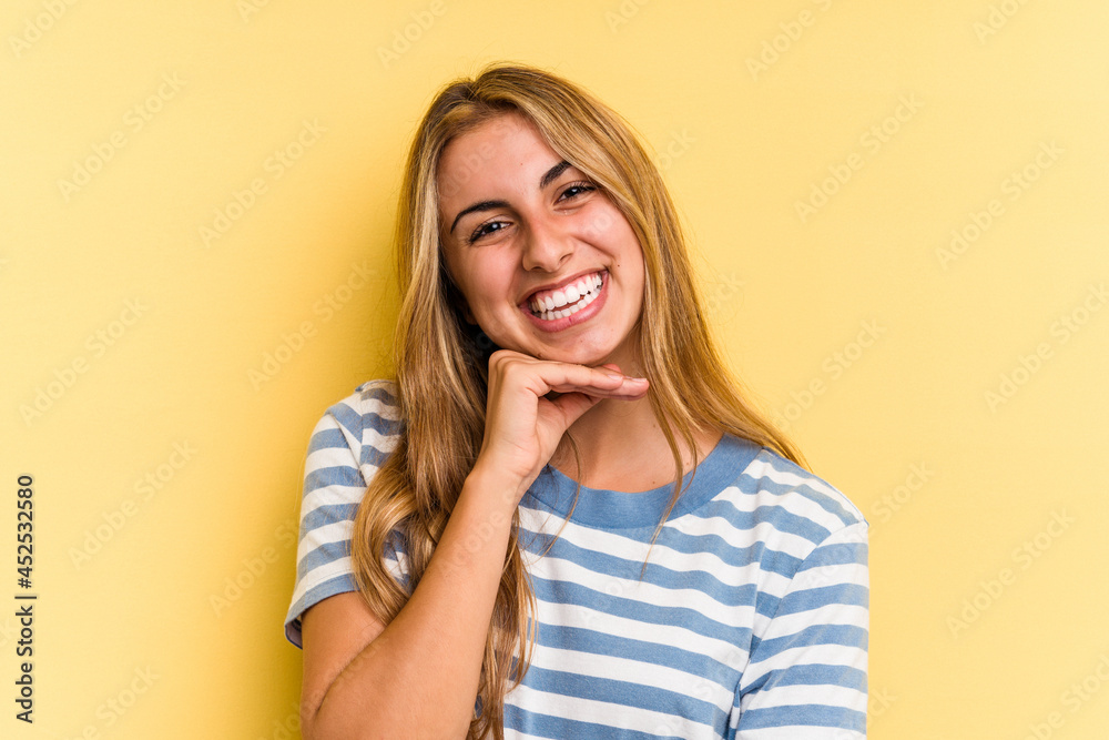 Young caucasian blonde woman isolated on yellow background smiling happy and confident, touching chin with hand. Young caucasian blonde woman isolated on yellow background smiling happy and confident, touching chin with hand.