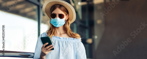 Young traveler in a medical protective mask on her face, a woman walks with suitcases and uses a mobile phone, over the airport building © Shopping King Louie