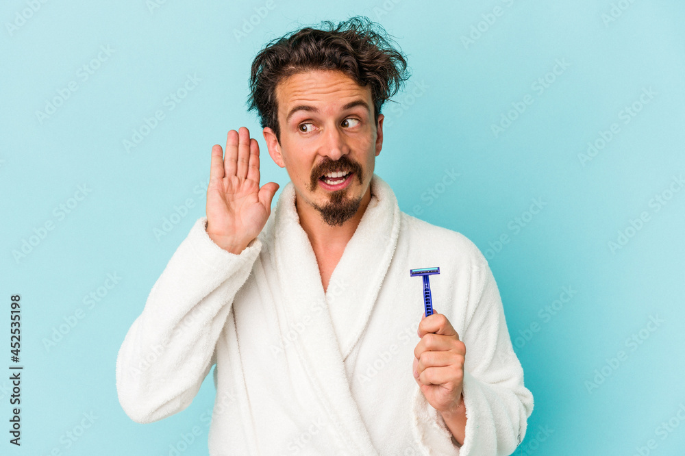 Young caucasian man holding a razor blade isolated on blue background trying to listening a gossip. Young caucasian man holding a razor blade isolated on blue background trying to listening a gossip.