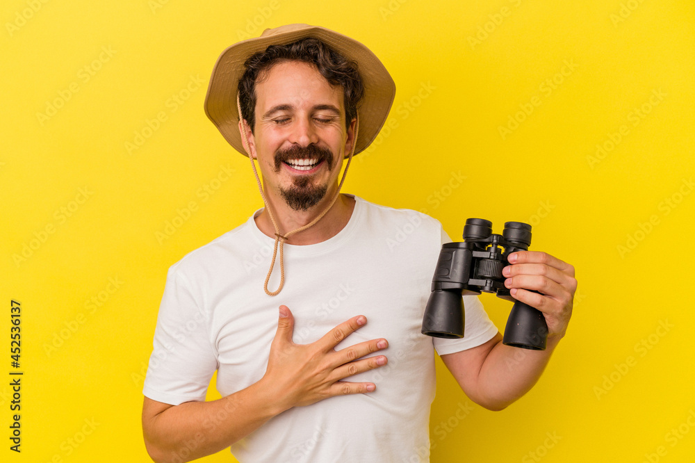 Young caucasian man holding binoculars isolated on yellow background laughs out loudly keeping hand on chest. Young caucasian man holding binoculars isolated on yellow background laughs out loudly keeping hand on chest.