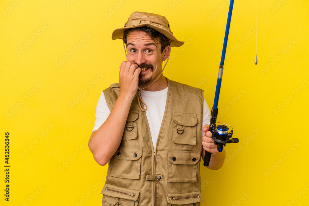 Young caucasian fisherman holding rod isolated on yellow background biting fingernails, nervous and very anxious. Young caucasian fisherman holding rod isolated on yellow background biting fingernails, nervous and very anxious.