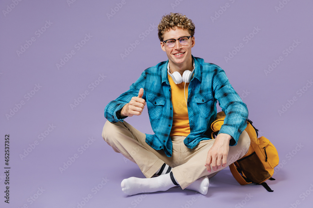 Full body young boy teen student wear casual clothes backpack headphones glasses sit crossed legs isolated on violet background studio portrait. Education in high school university college concept Full body young boy teen student wear casual clothes backpack headphones glasses sit crossed legs isolated on violet background studio portrait. Education in high school university college concept