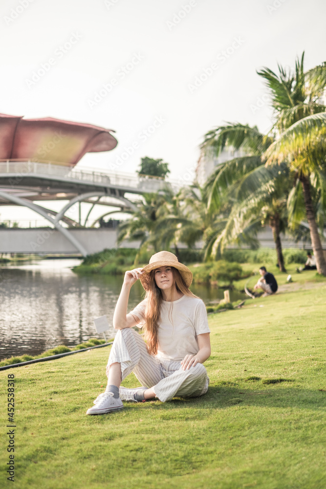 Young beautiful blonde woman wearing white casual clothes, straw hat and sneakers sitting on the grass in the park. Trendy casual summer outfit.
