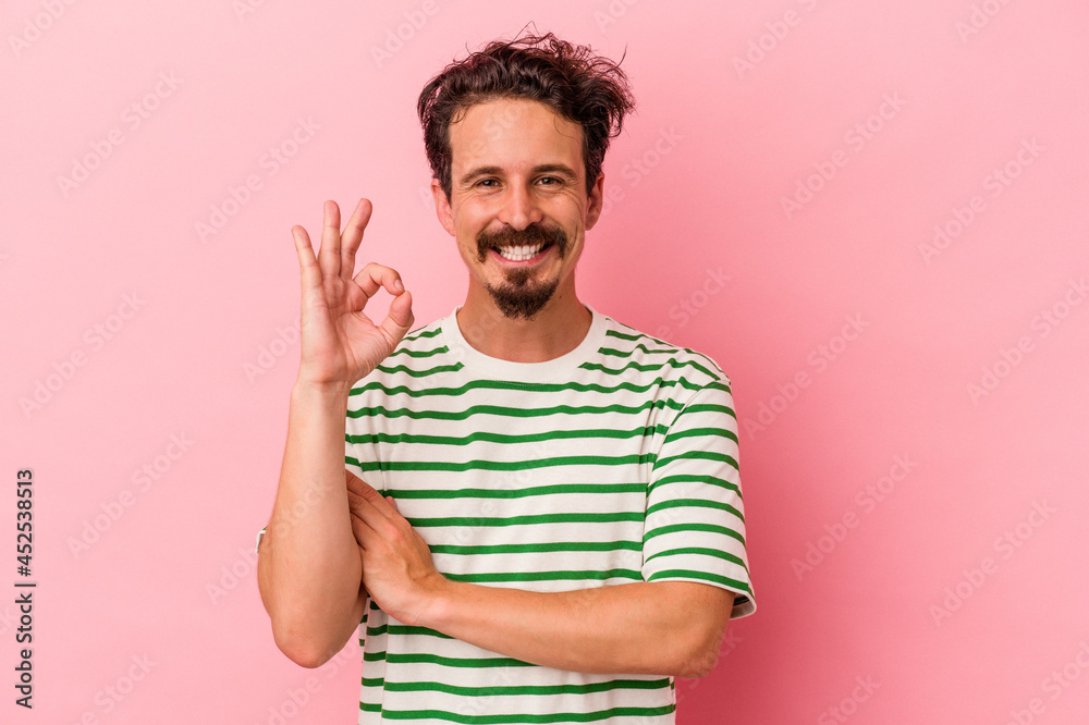 Young caucasian man isolated on pink background winks an eye and holds an okay gesture with hand. Young caucasian man isolated on pink background winks an eye and holds an okay gesture with hand.