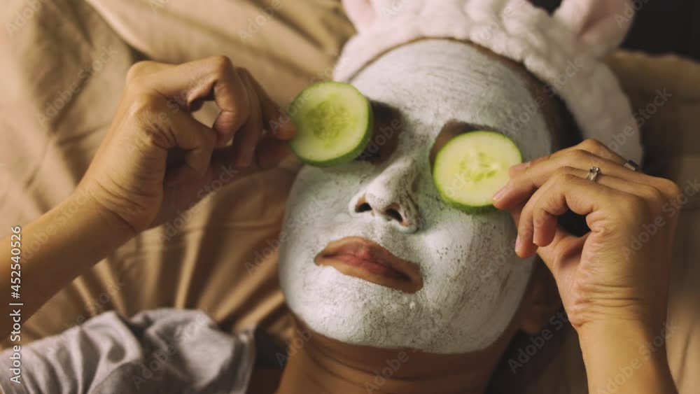 Face focusing authentic action of medium age asian woman who is placing sliced cucumber on her eyes with masking face with mud or clay for moisturizing and beauty skin on bed with relaxing warm light.
