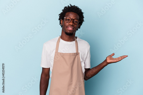 Valokuvatapetti Young african american store clerk isolated on blue background showing a copy space on a palm and holding another hand on waist