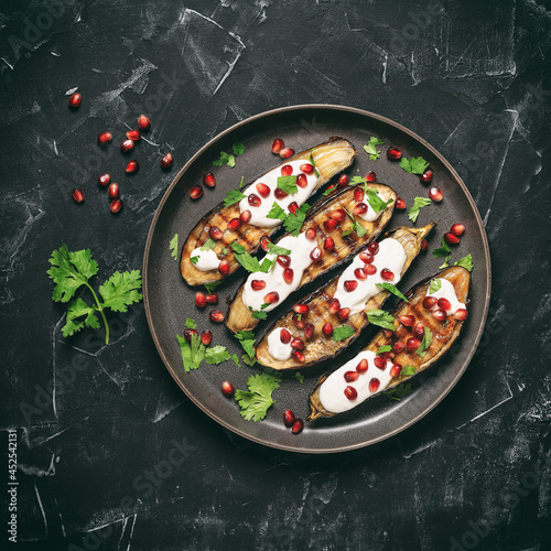 Grilled eggplants with yoghurt sauce and pomegranate on a black stone background. Top view, flat lay. Square toned photo.