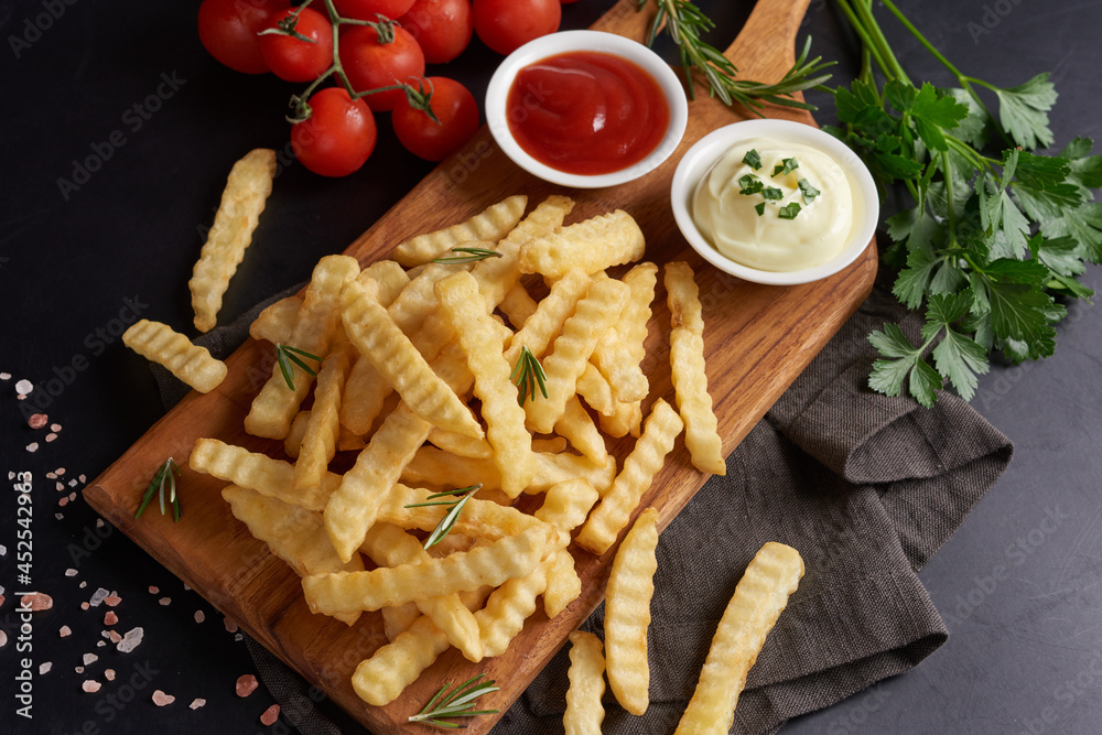 Homemade baked potato fries with mayonnaise, tomato sauce and rosemary on wooden board. tasty french fries on cutting board, in brown paper bag on black stone table background, unhealthy food. Homemade baked potato fries with mayonnaise, tomato sauce and rosemary on wooden board. tasty french fries on cutting board, in brown paper bag on black stone table background, unhealthy food.