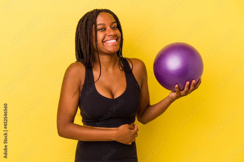 Young african american woman holding a pilates ball isolated on yellow background laughing and having fun. Young african american woman holding a pilates ball isolated on yellow background laughing and having fun.