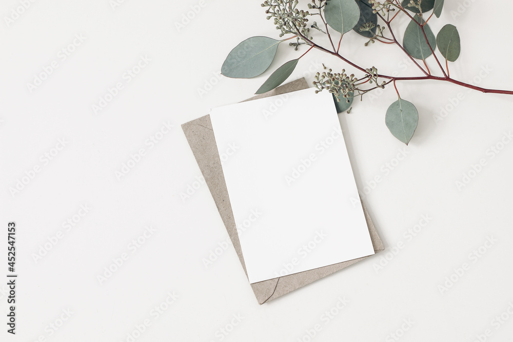 Feminine wedding, birthday stationery composition. Blank greeting card, invitation mockup with craft envelope. Green Eucalyptus populus branches isolated on table background. Flat lay, top view. Feminine wedding, birthday stationery composition. Blank greeting card, invitation mockup with craft envelope. Green Eucalyptus populus branches isolated on table background. Flat lay, top view.