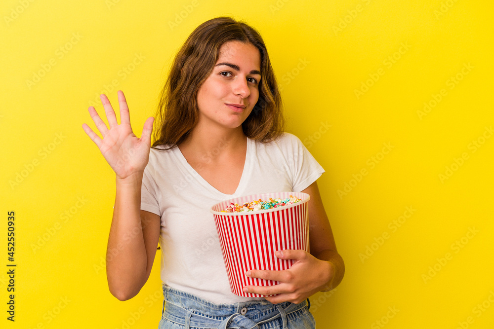 Young caucasian woman holding a popcorns isolated on yellow background  smiling cheerful showing number five with fingers.