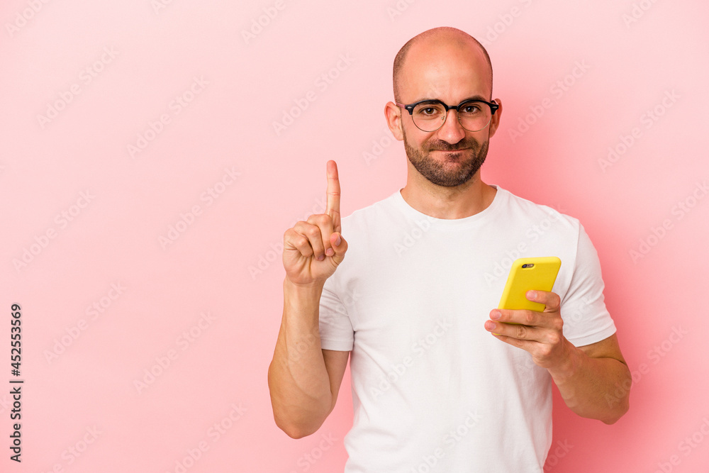 Young caucasian bald man holding mobile phone isolated on pink background showing number one with finger. Young caucasian bald man holding mobile phone isolated on pink background showing number one with finger.