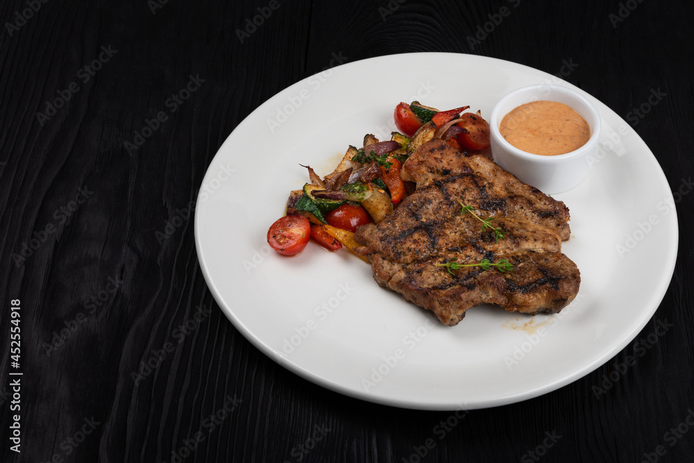 Fried pork chop with grilled vegetables and sauce on white plate on black wooden background