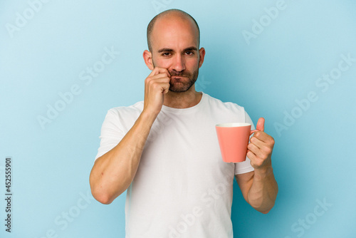 Young bald man holding a mug isolated on blue background  with fingers on lips keeping a secret. © Asier