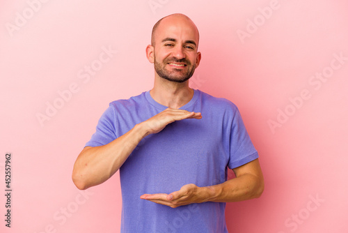 Young caucasian bald man isolated on pink background holding something with both hands, product presentation.