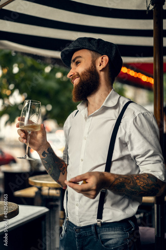 Young bearded man in a cafe on the street with a glass of wine. Romantic guy in a white shirt cap and suspenders in the city. Peaky Blinders. old fassion retro. photo