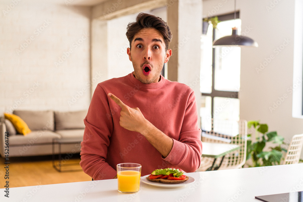 Young mixed race man having breakfast in his kitchen pointing to the side Young mixed race man having breakfast in his kitchen pointing to the side