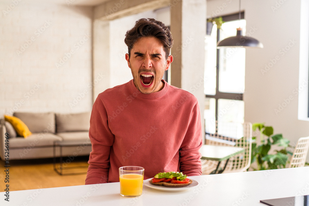 Young mixed race man having breakfast in his kitchen screaming very angry and aggressive. Young mixed race man having breakfast in his kitchen screaming very angry and aggressive.