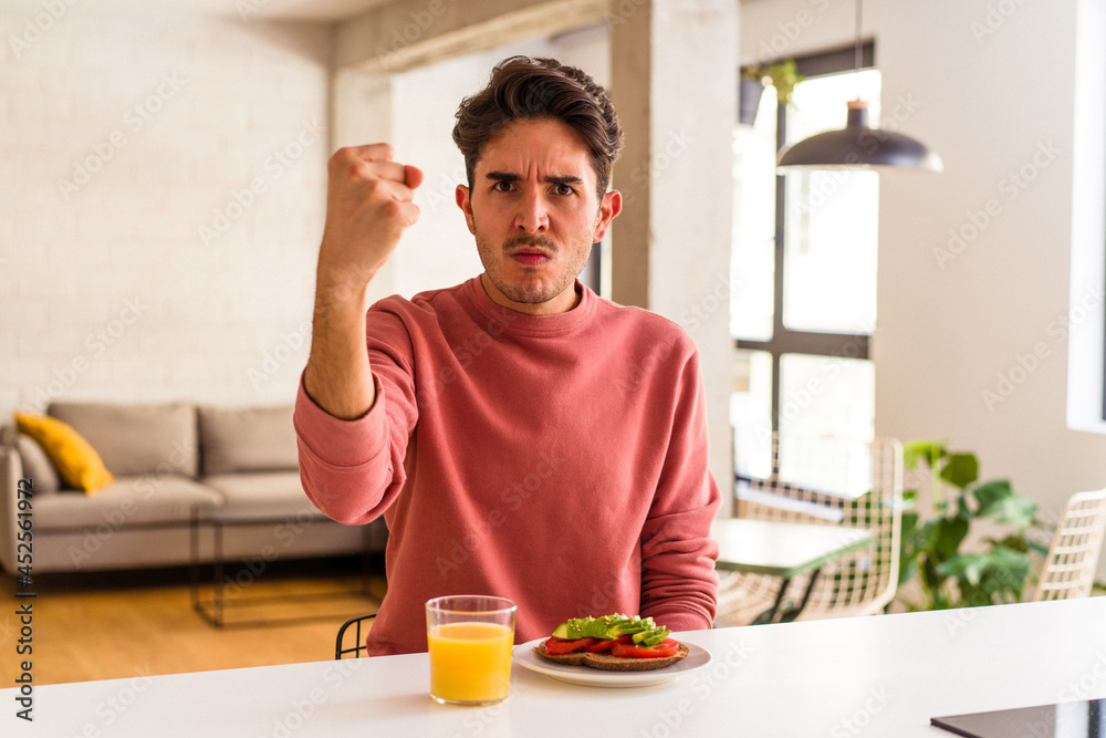 Young mixed race man having breakfast in his kitchen showing fist to camera, aggressive facial expression. Young mixed race man having breakfast in his kitchen showing fist to camera, aggressive facial expression.