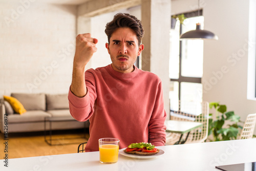 Young mixed race man having breakfast in his kitchen showing fist to camera, aggressive facial expression.