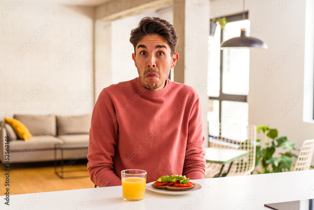 Young mixed race man having breakfast in his kitchen shrugs shoulders and open eyes confused.