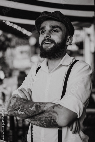 Young bearded tattooed man in a cafe on the street with a glass of wine. A romantic guy in a white cap and suspenders in the city. Peaky Blinders. old fashionable retro.