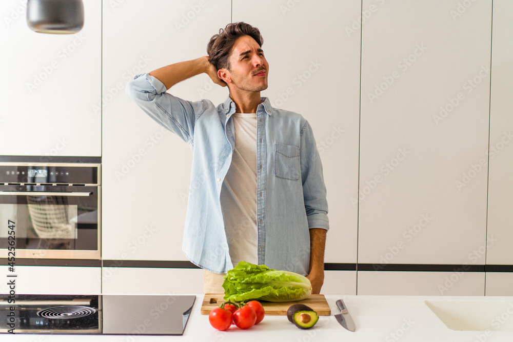Young mixed race man preparing a salad for lunch touching back of head, thinking and making a choice.