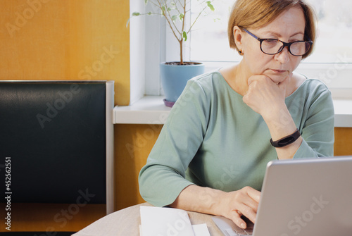 Concentrated senior woman working on laptop sitting at home indoor near window. Listening audio course, learning language, watching online webinar,shopping, booking using wireless internet connection