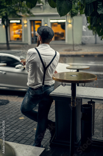 Young bearded tattooed man in a cafe on the street with a glass of wine. A romantic guy in a white cap and suspenders in the city. Peaky Blinders. old fashionable retro.