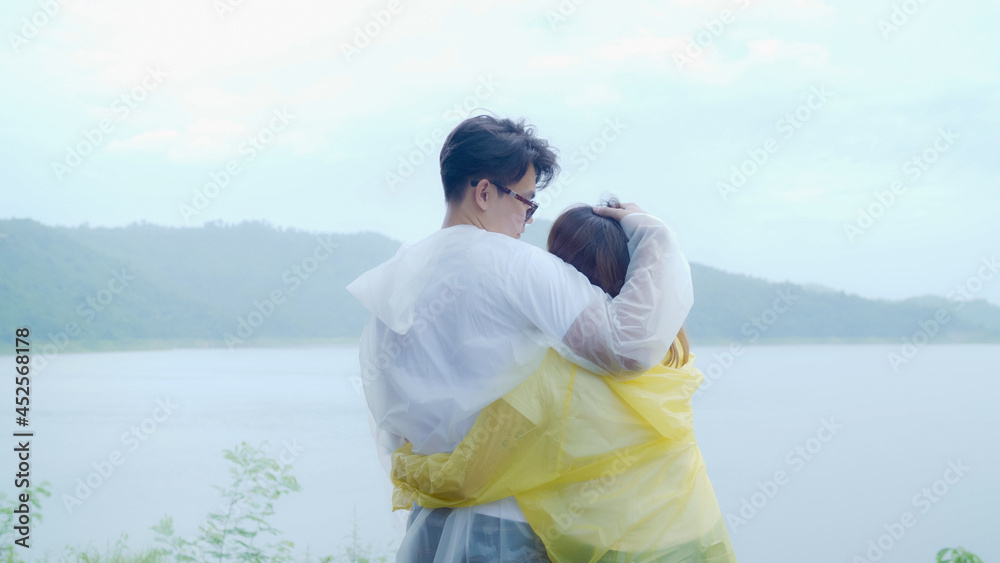 Young Asian sweet couple feeling happy using romantic time playing rain while wearing raincoat standing near lake. Lifestyle couple enjoy and relax in rainy day. Young Asian sweet couple feeling happy using romantic time playing rain while wearing raincoat standing near lake. Lifestyle couple enjoy and relax in rainy day.