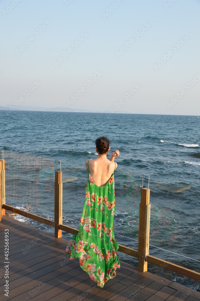 Beautiful woman wearing a green flower maxi dress, standing on the edge of a bar on rock island Phu Quoc, Vietnam, looking over the ocean.