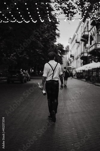 Young bearded tattooed man drinking wine from a glass on the street. Romantic guy in a white shirt, cap and suspenders in the city. Peaky Blinders. old-fashioned, retro.