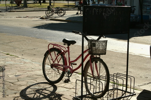 Bicicleta rosa con canasto estacionada con sol de frente y sombras oscuras con fondo de calle y plaza. photo