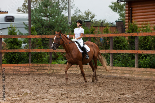 A girl in white equestrian clothes and wearing a helmet, riding a brown horse