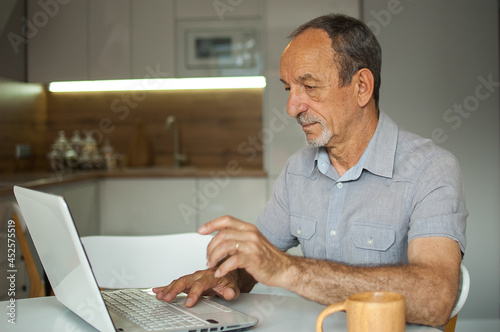 Trendy mature man is working from home with laptop sitting at the table in his kitchen, happy retirement, home-office concepts