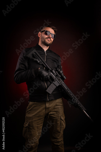 A man in dark clothes holds an automatic carbine in his hands. Eyes covered with sunglasses, hands are in gloves. A figure with a weapon on a dark background with a red light spot.