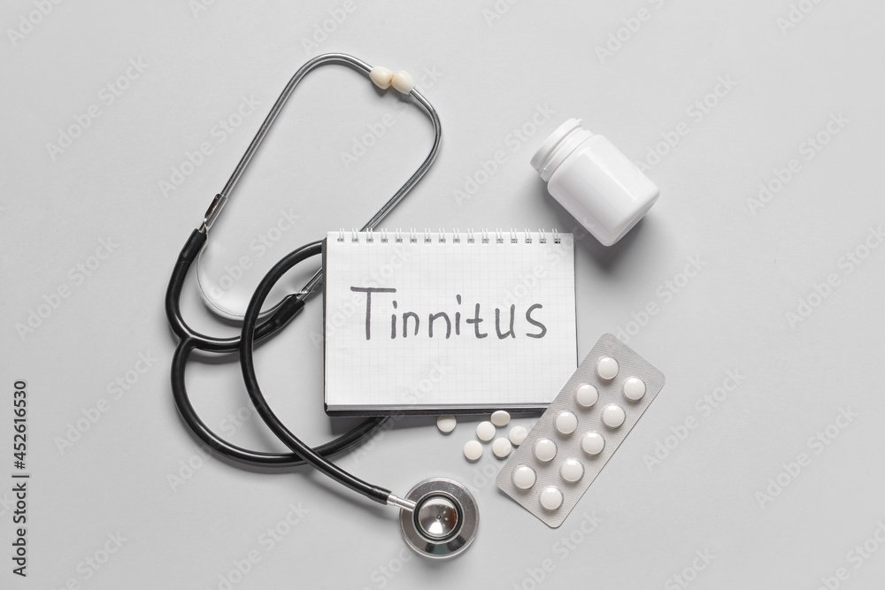 Notebook with word TINNITUS, stethoscope and pills on light background Notebook with word TINNITUS, stethoscope and pills on light background