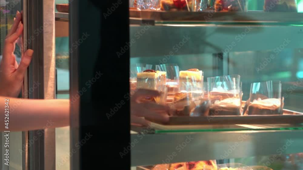 Asian woman coffee shop employee placing pastry and cake in bakery refrigerator showcase at cafe. Female waitress preparing restaurant for service to customer. Small business and part time job concept Asian woman coffee shop employee placing pastry and cake in bakery refrigerator showcase at cafe. Female waitress preparing restaurant for service to customer. Small business and part time job concept