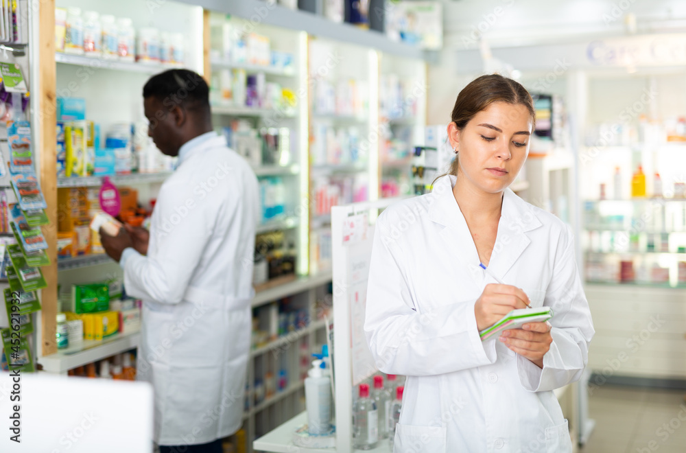 Female pharmacist standing in salesroom of drugstore with notebook in hands. Her colleague working behind.