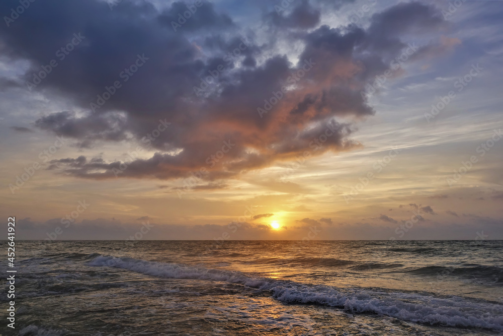 Dawn over the Caribbean Sea. The sun is low on the horizon. There are mauve clouds in the sky. The surf waves are foaming on the sandy beach. A sunny path on the water. Mexico