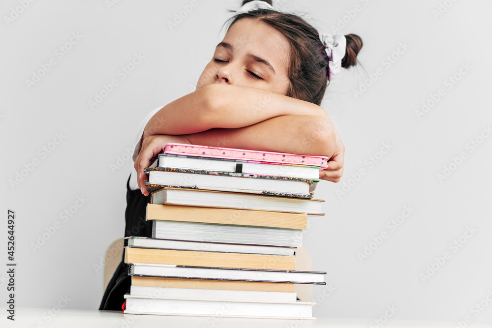Bored and tired schoolgirl sleeping on a pile of books on the desk against light grey background. Exhausted little girl learner falling asleep during reading lot of books. Consuming education concept. Bored and tired schoolgirl sleeping on a pile of books on the desk against light grey background. Exhausted little girl learner falling asleep during reading lot of books. Consuming education concept.