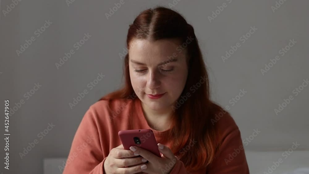 Phone. Young caucasian millennial girl using smartphone. Young girl holding cellphone, chatting in social networks, sitting on bed in living room.