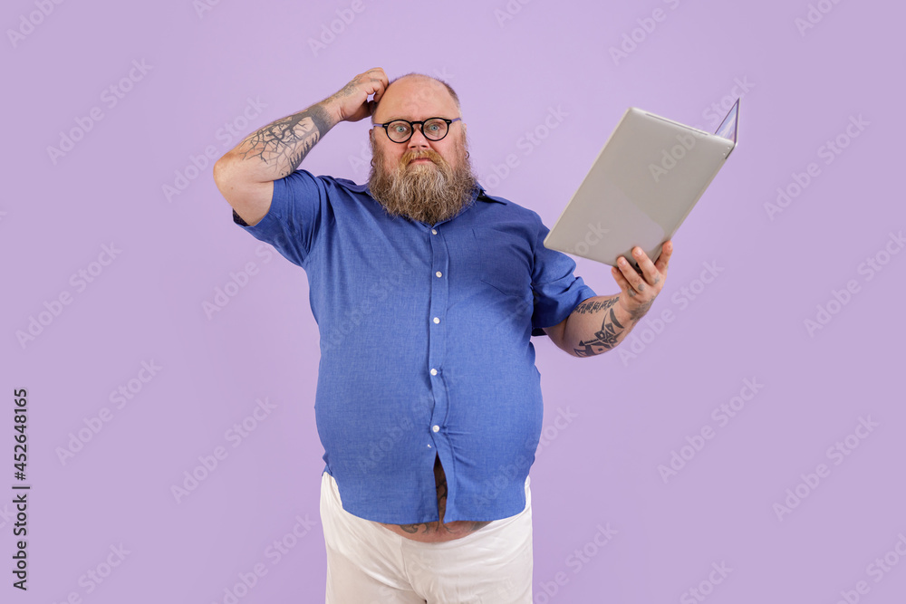 Doubting middle aged man with overweight in tight blue shirt scratches head holding laptop as book on purple background in studio Doubting middle aged man with overweight in tight blue shirt scratches head holding laptop as book on purple background in studio