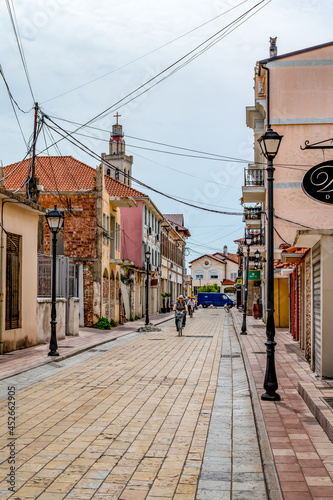 Shkoder, Albania - June 21, 2021: Cyclists ride on Rruga G'juhadol pedestrian street in Shkoder, vertical. Ancient architecture and many electric wires on the city street of the Albanian town photo