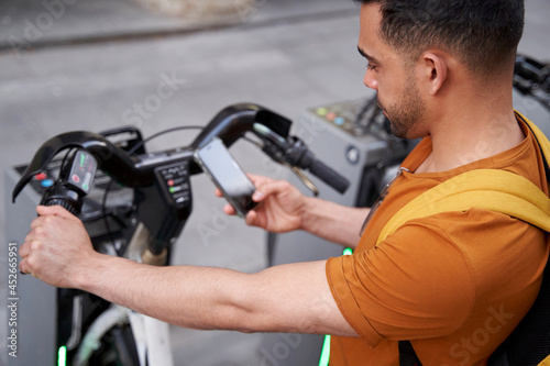 Young Latino man stands by the parking lot with a rental bike and uses a smartphone with a smile on his face. Eco-friendly transport concept