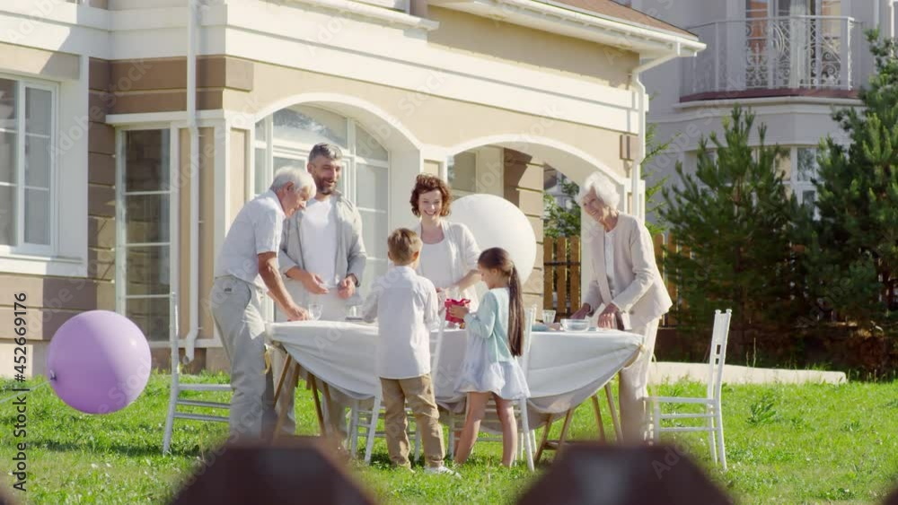 Happy multigenerational family having holiday dinner outdoor on summer day: parents and grandparents smiling and setting table while little boy giving birthday present to sister Happy multigenerational family having holiday dinner outdoor on summer day: parents and grandparents smiling and setting table while little boy giving birthday present to sister