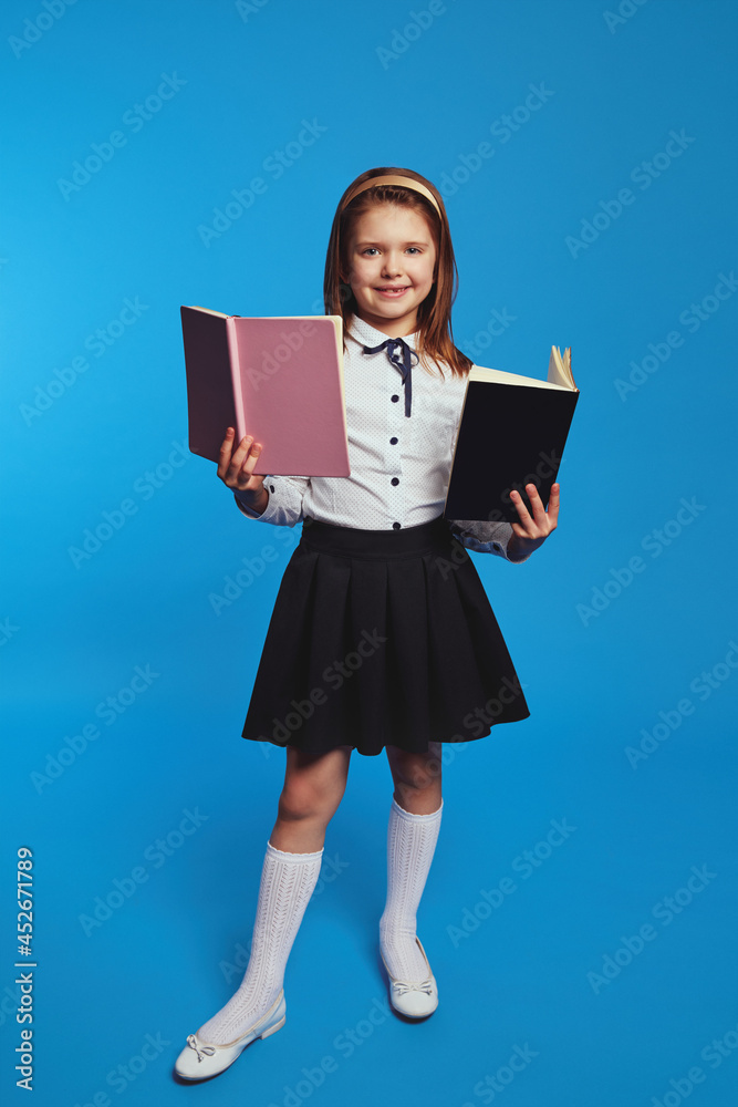 Vertical photo of full body adorable kid wears school uniform, holding  textbooks in hands, poses over blue background. Girl learns from textbook  Photos | Adobe Stock